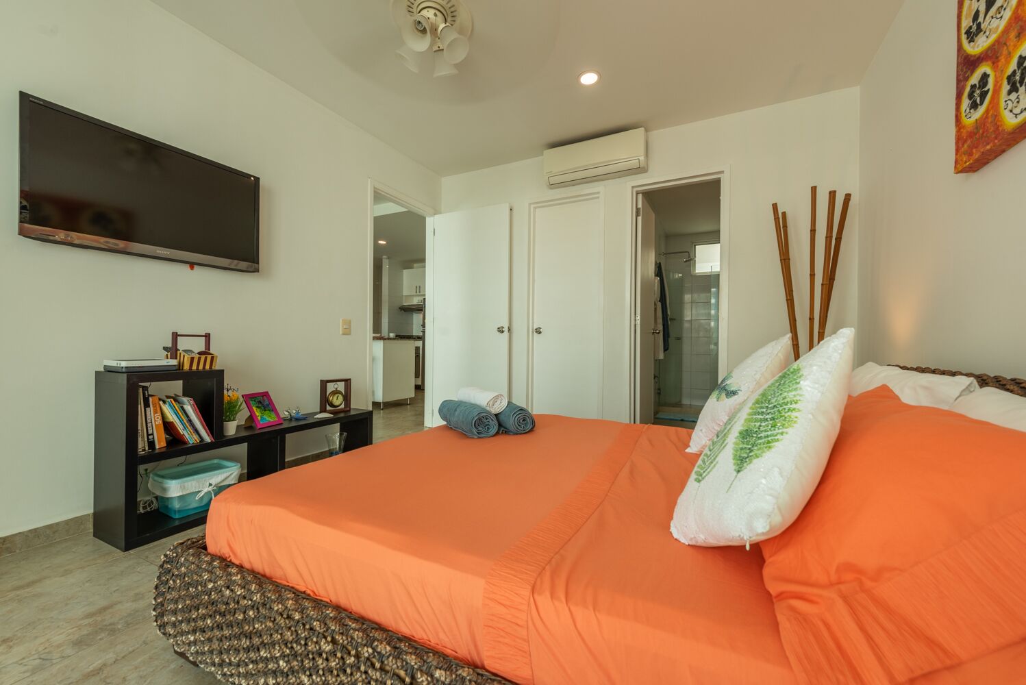 Un dormitorio con colcha naranja, un televisor de pantalla plana, una estantería y una puerta que da al baño.