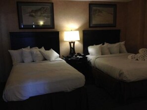 Two beds with dark headboards and multiple pillows, a bedside table with a lamp and personal items, and framed artwork adorn the room.