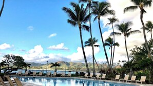 A pool area with lounge chairs and palm trees, surrounded by a stone-tiled deck and a view of a body of water and hills. - Beautiful condo,air conditioning in bedrooms,ocean 100 yards from unit,sleeps 4 (Kapaa)