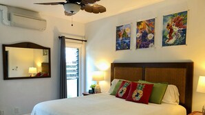 A hotel room with a large bed, a ceiling fan, a window with curtains, a wall-mounted air conditioner, and three paintings on the wall. - Beautiful condo,air conditioning in bedrooms,ocean 100 yards from unit,sleeps 4 (Kapaa)
