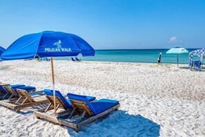 A beach with blue umbrellas labeled "PELICAN WALK," wooden lounge chairs, and a clear view of the ocean.