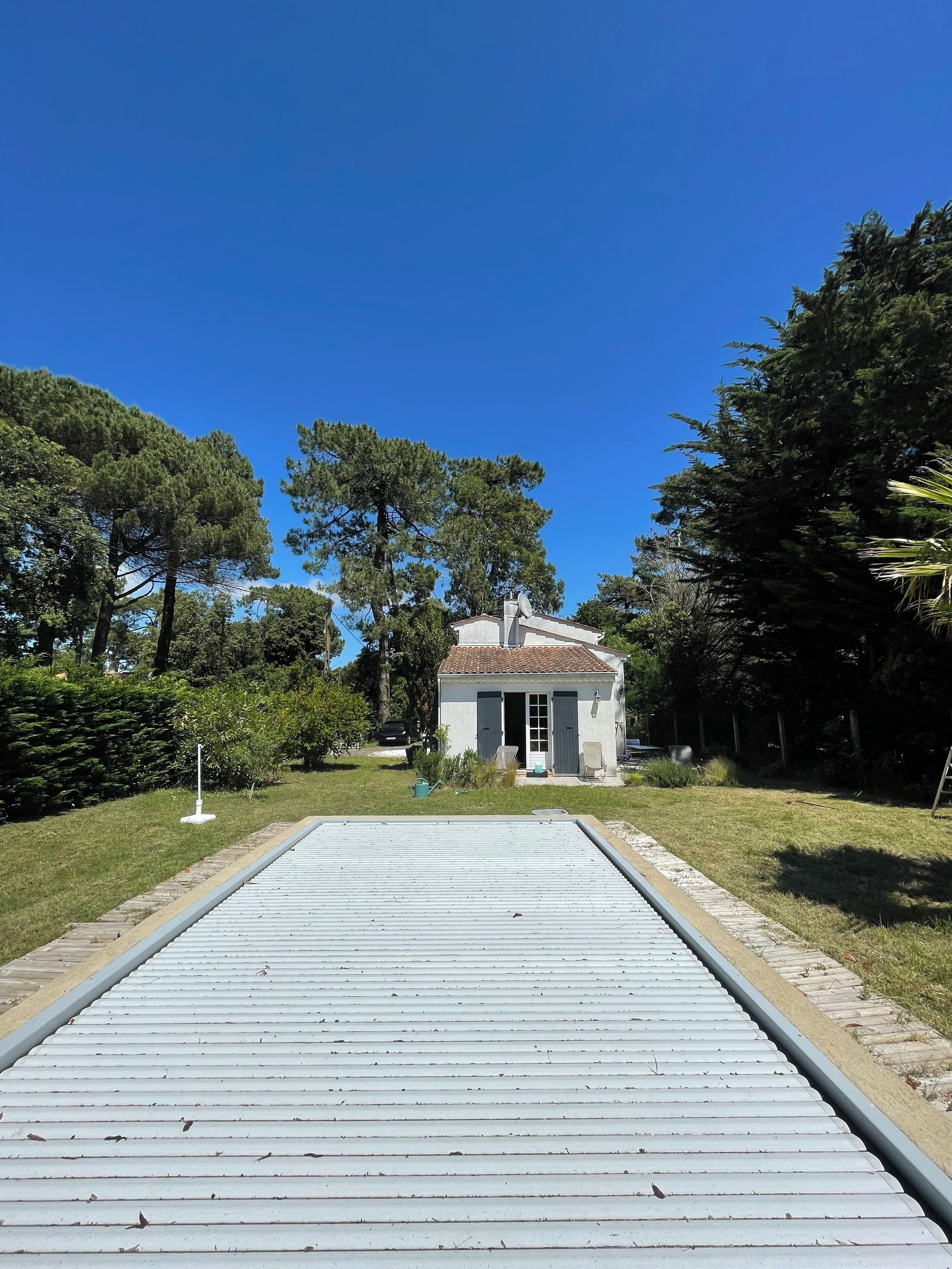 Un chemin menant à une maison avec un toit en tuiles et une antenne parabolique, entourée d’arbres et d’une pelouse.