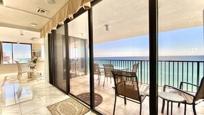 A balcony with a view of the sea, a dining area with chairs and a table, and a large window with curtains.
