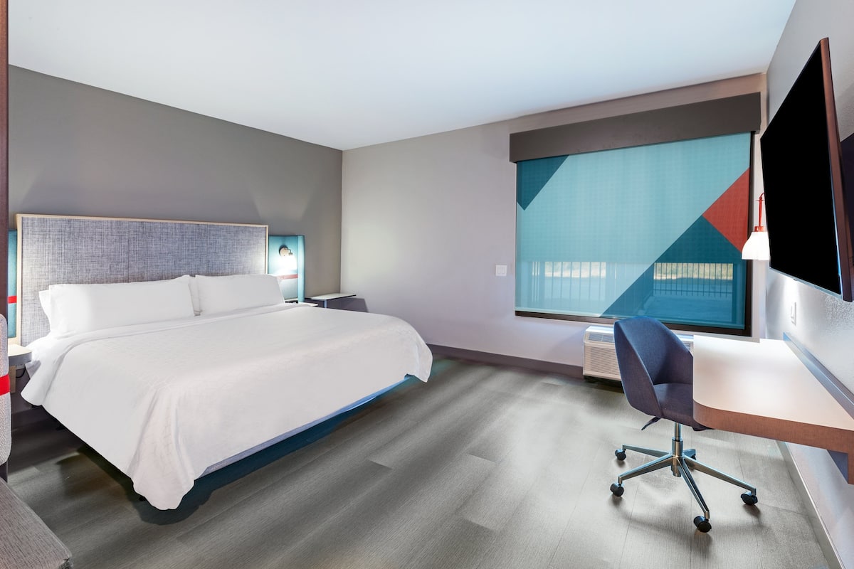 Une chambre d’hôtel moderne dotée d’un grand lit, d’un bureau avec une chaise, d’un téléviseur à écran plat et d’une grande fenêtre offrant une vue géométrique.