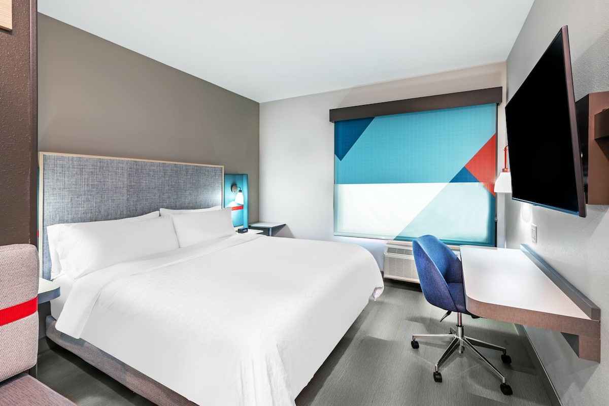 Une chambre d’hôtel moderne dotée d’un grand lit, d’un bureau, d’une chaise, d’une télévision et d’une fenêtre offrant une vue géométrique.