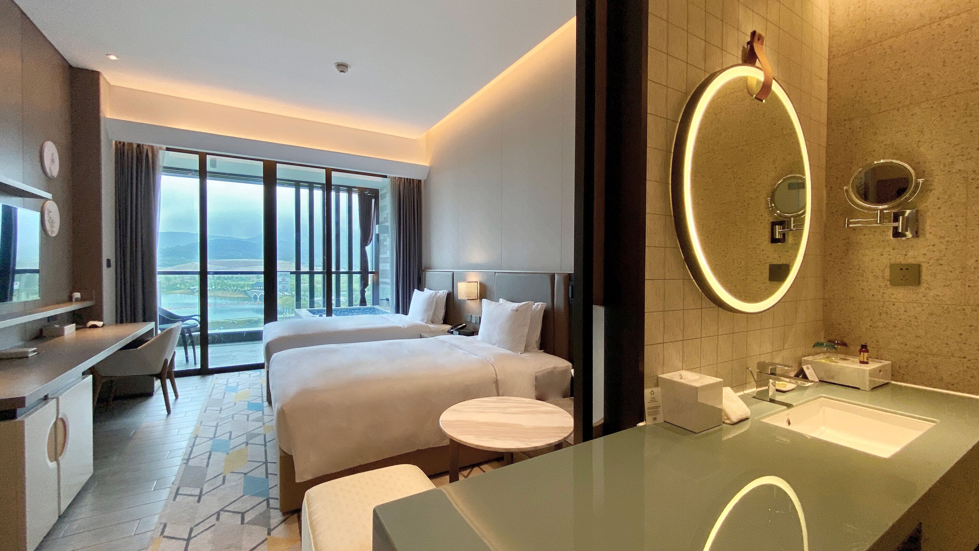 A hotel room with a bed, a desk, a round mirror, and a bathroom with a sink and mirror.