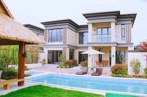 A modern house with a swimming pool, lounge chairs, and a thatched roof structure.