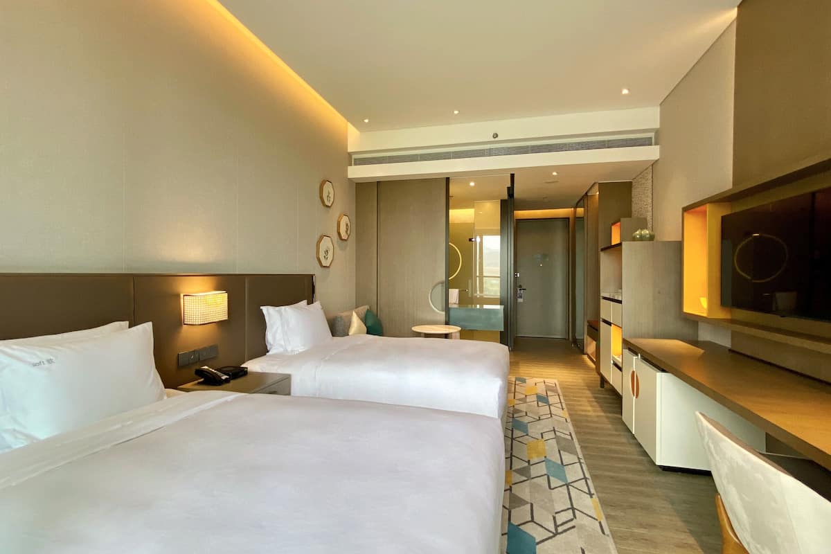 Habitación de hotel con dos camas, televisión, escritorio y baño visible a través de una puerta abierta.