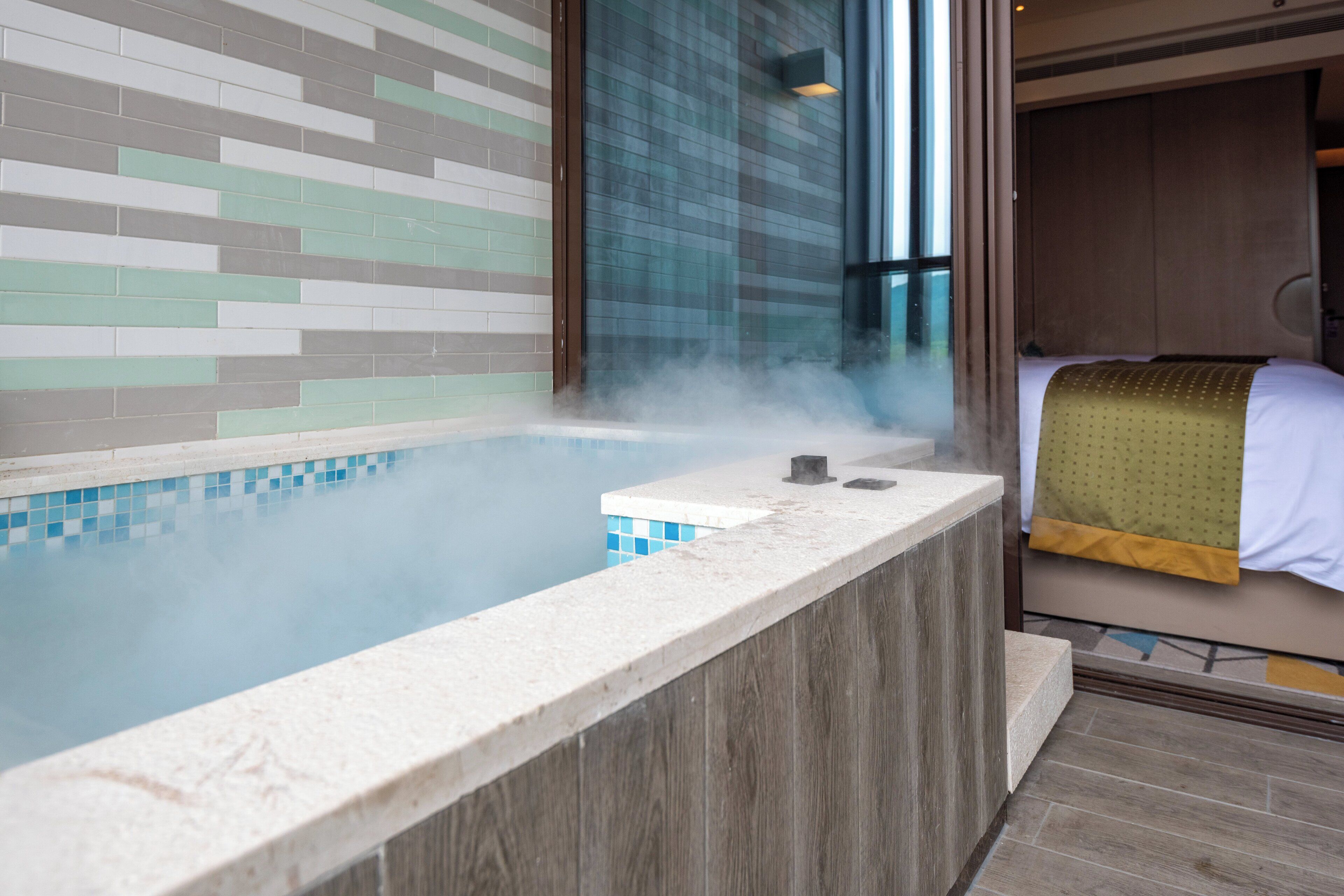 A steam room with a tiled wall, a wooden bench, and a bed with a green blanket.