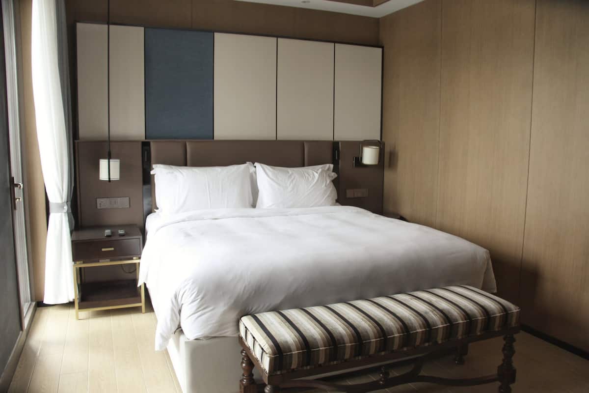Una cama bien tendida con sábanas blancas, una mesita de noche de madera con una lámpara, y un banco a rayas.