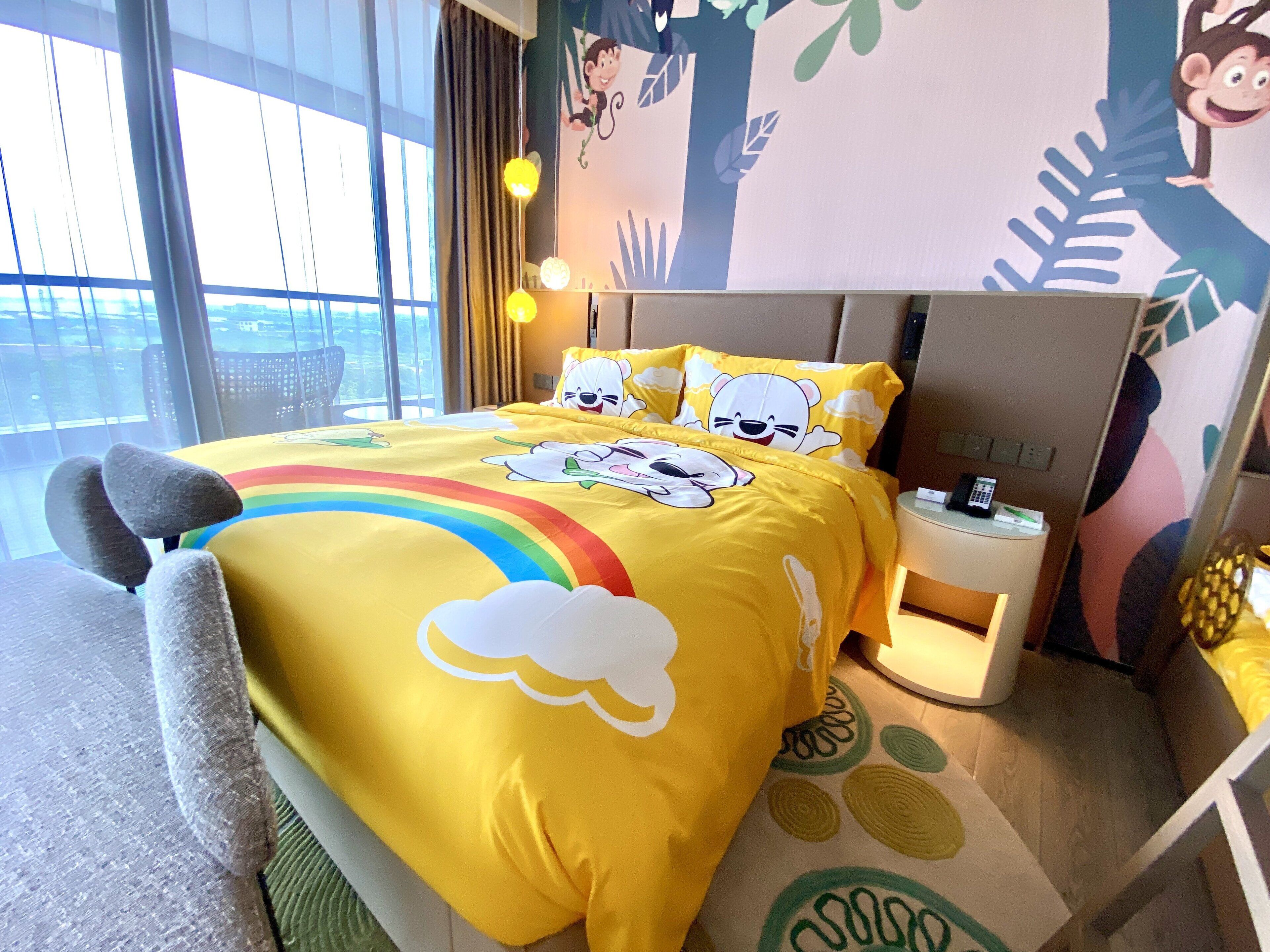 Hotelkamer met een bed met tekenfilmfiguren en een regenboogdesign, een nachtkastje met een telefoon en een lamp, en uitzicht op de stad door grote ramen.