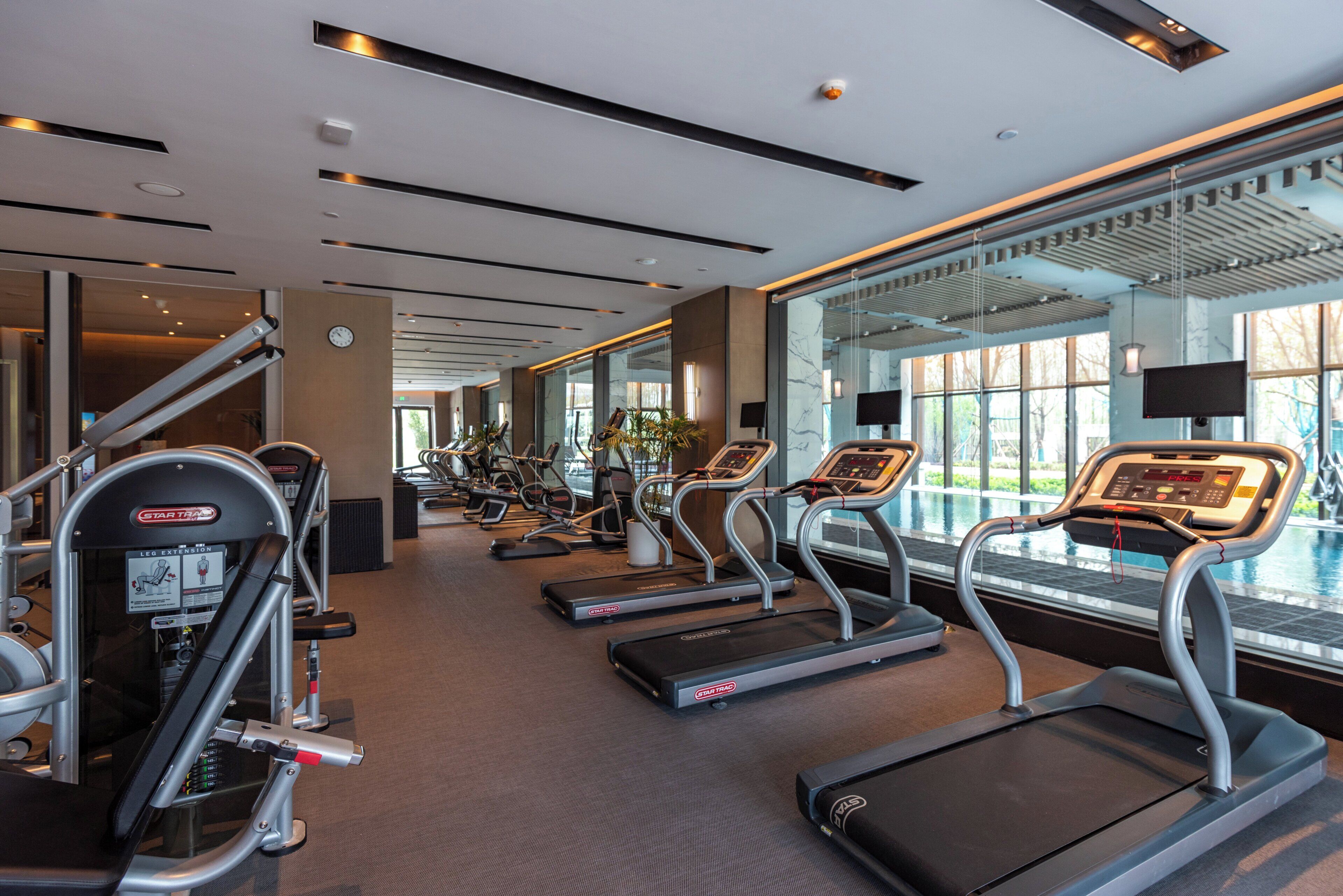A modern gym with treadmills, leg extension machines, and a large window overlooking a pool.