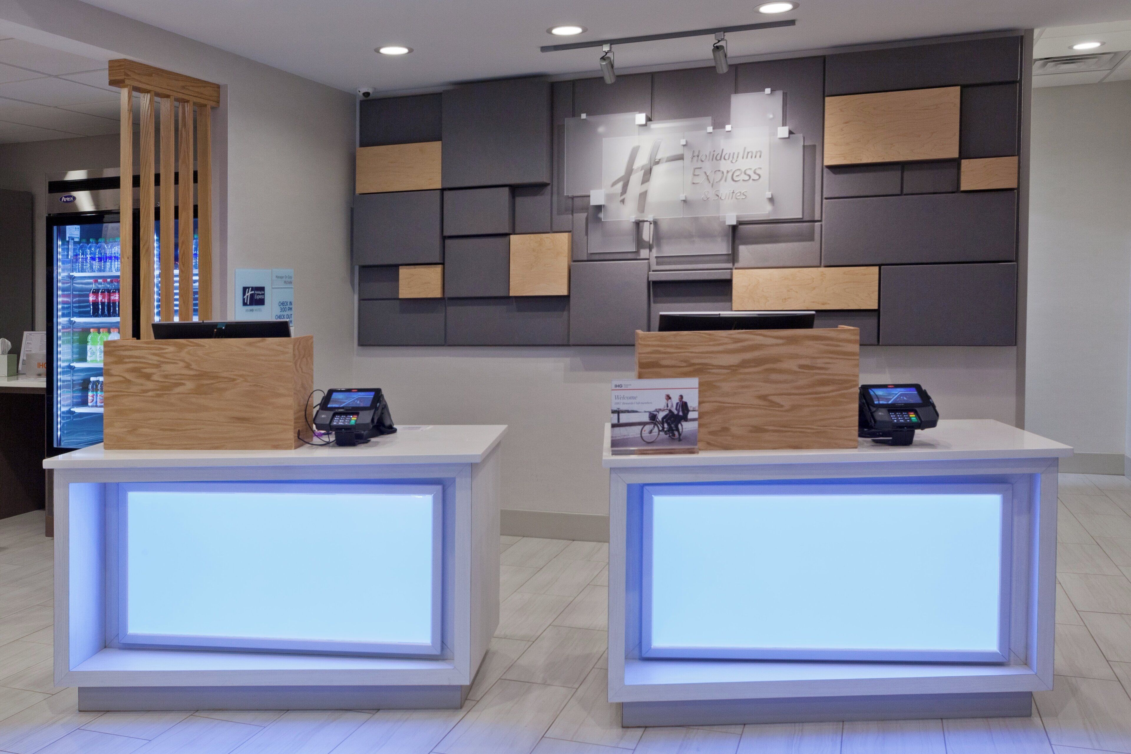 A reception desk with a digital display, a refrigerator with beverages, and a wall with a sign for holiday inn express suites.