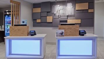 A reception desk with a digital display, a refrigerator with beverages, and a wall with a sign for holiday inn express suites.