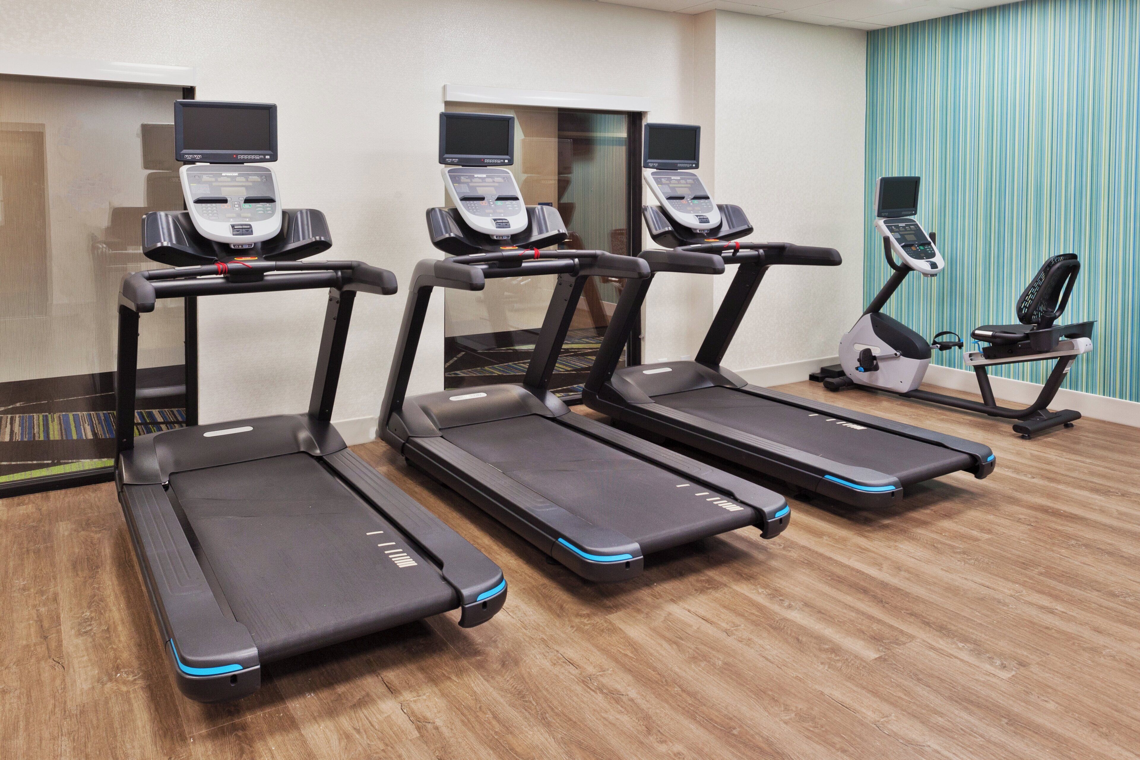 Quatre tapis de course dans une pièce dont le sol est en bois et un des murs est décoré de rayures bleues.