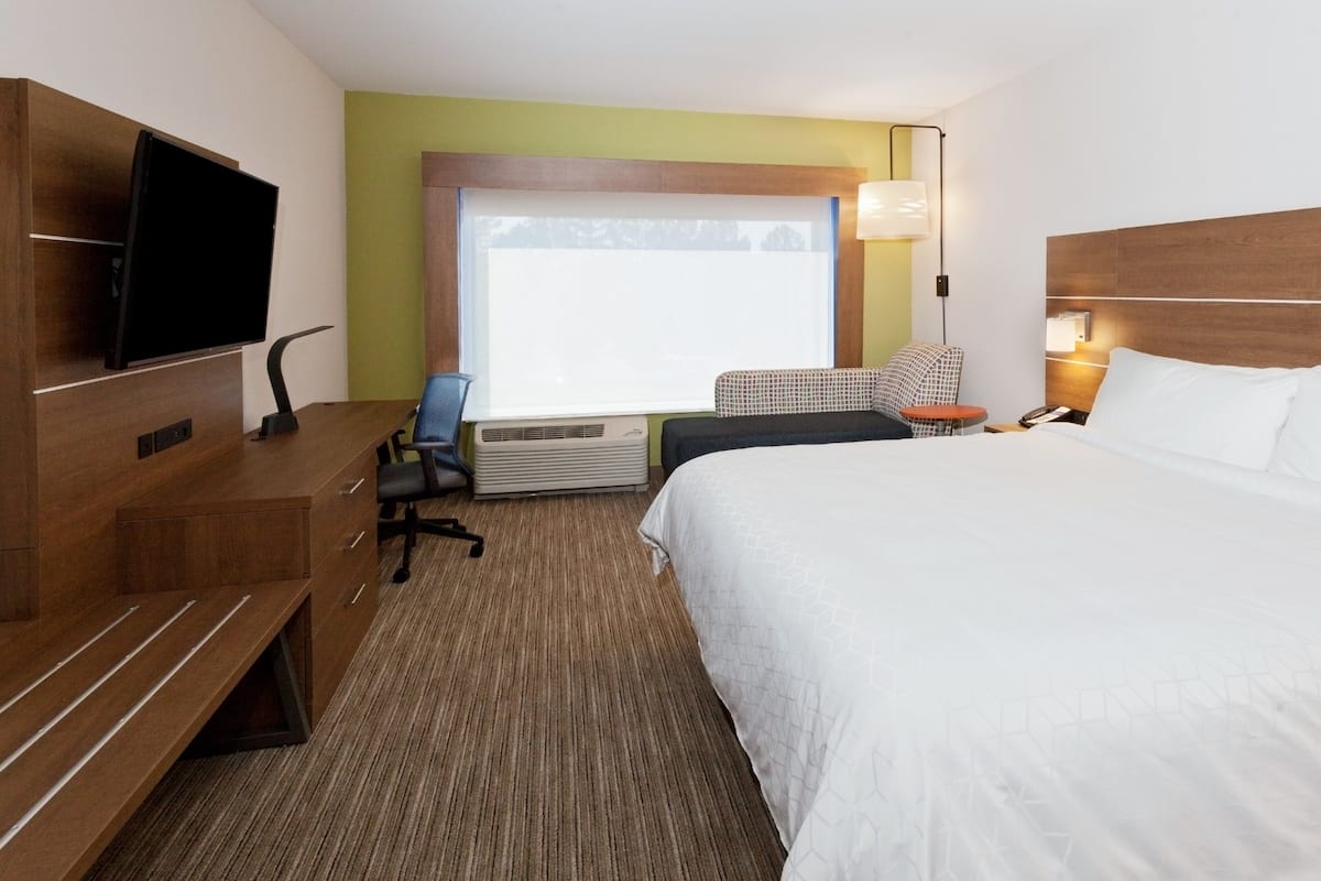 Una habitación de hotel con cama, escritorio con televisor de pantalla plana, una silla, una mesita y ventana con persianas.