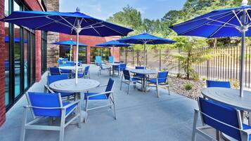 An outdoor patio area with round tables and blue chairs, shaded by large umbrellas.