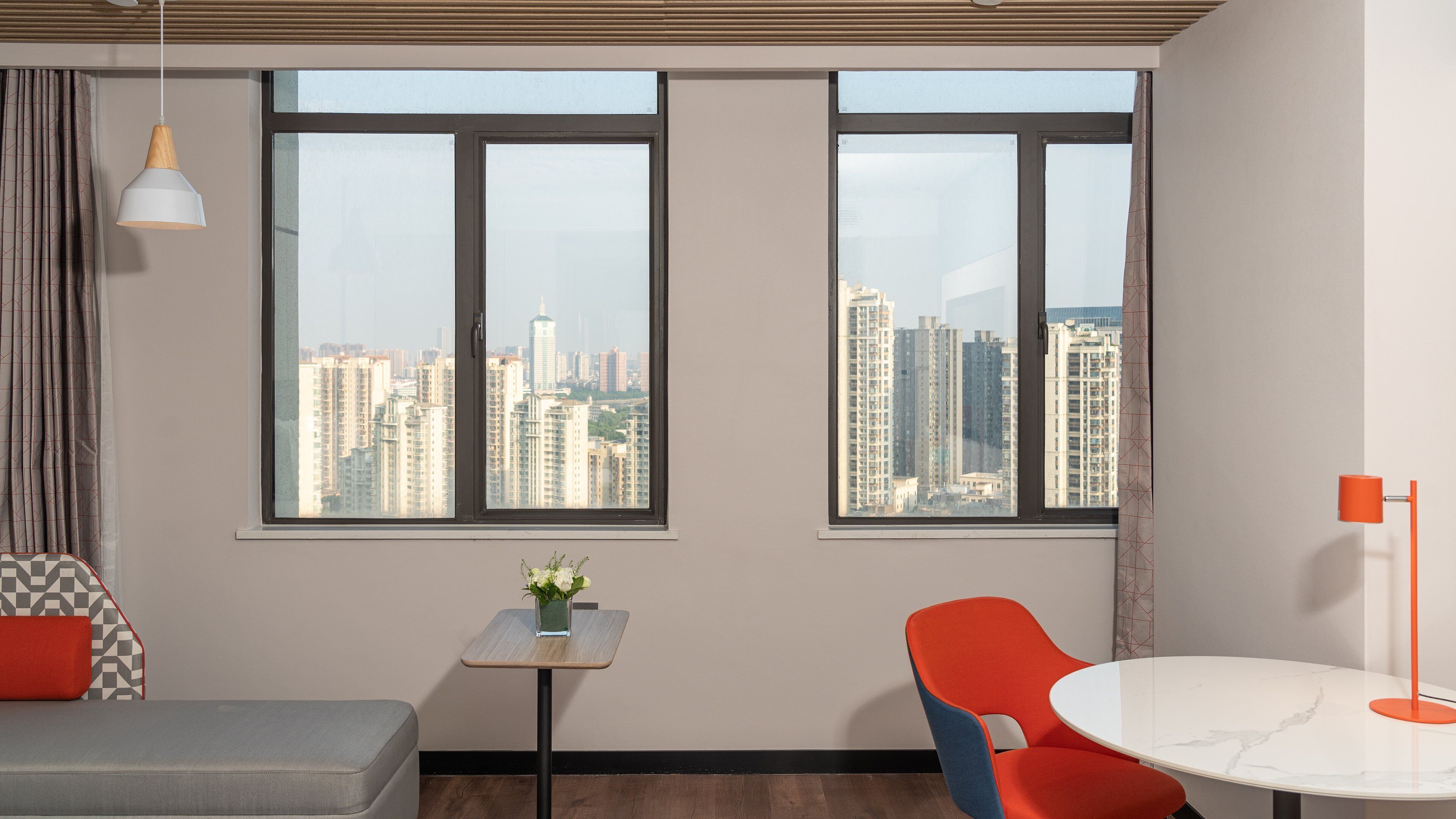 A modern room with two windows, a small table with a plant, a red chair, and a grey sofa.