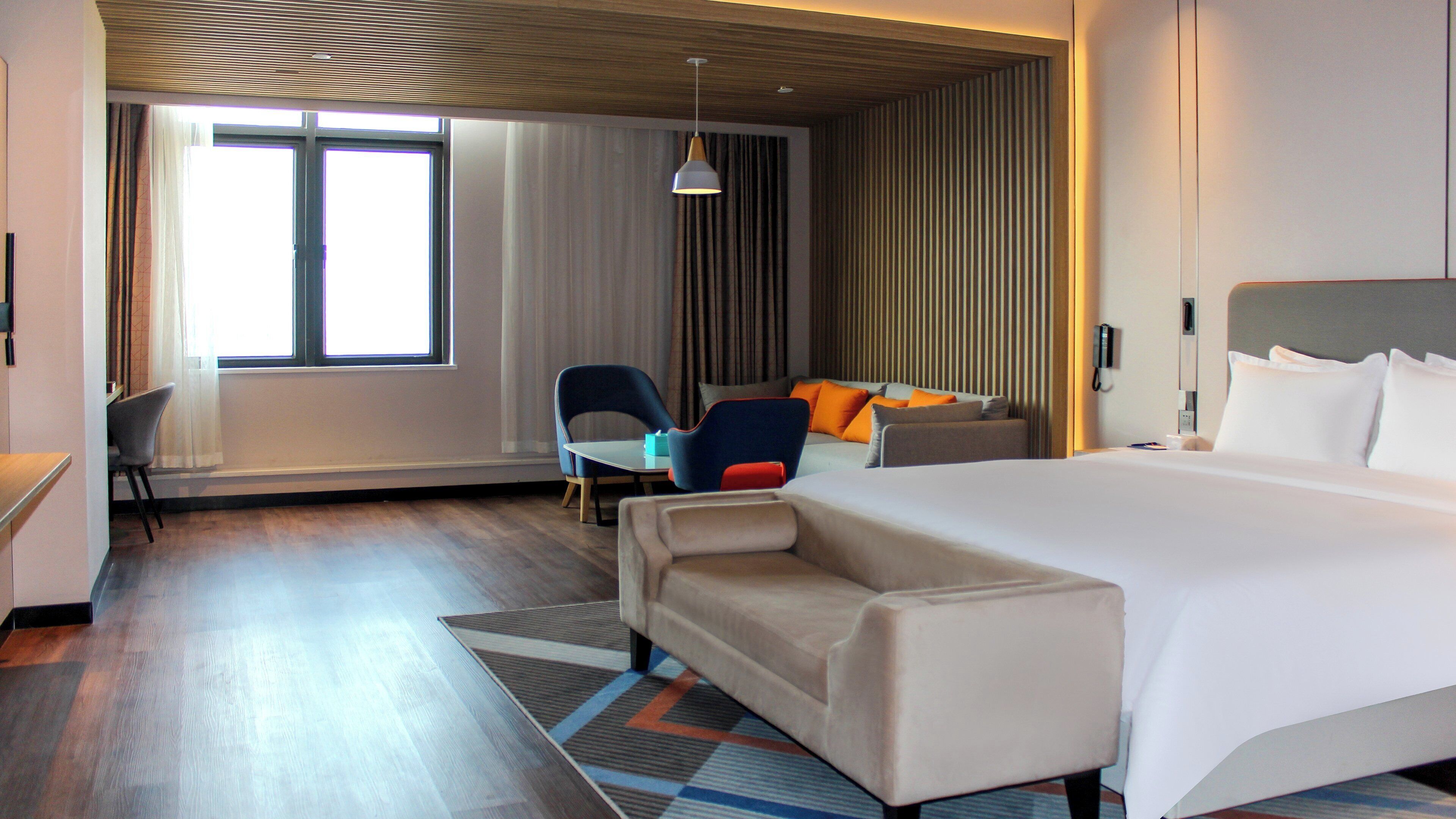 A hotel room with a large bed, a sofa, a desk, and a chair. There is a window with curtains and a hanging light fixture.