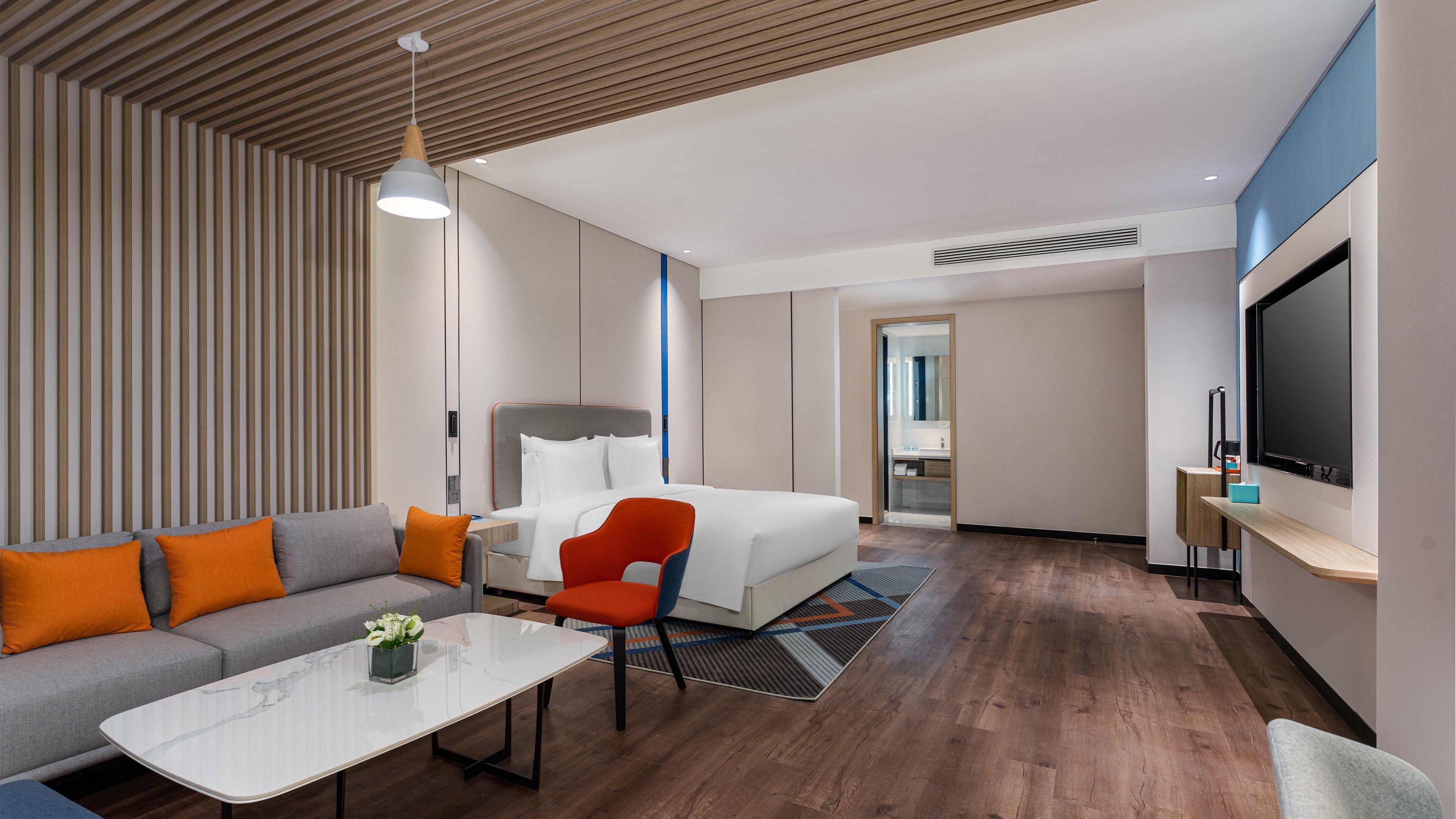 A modern hotel room with a sofa, a red chair, a bed, a TV, and a bathroom visible through an open door.