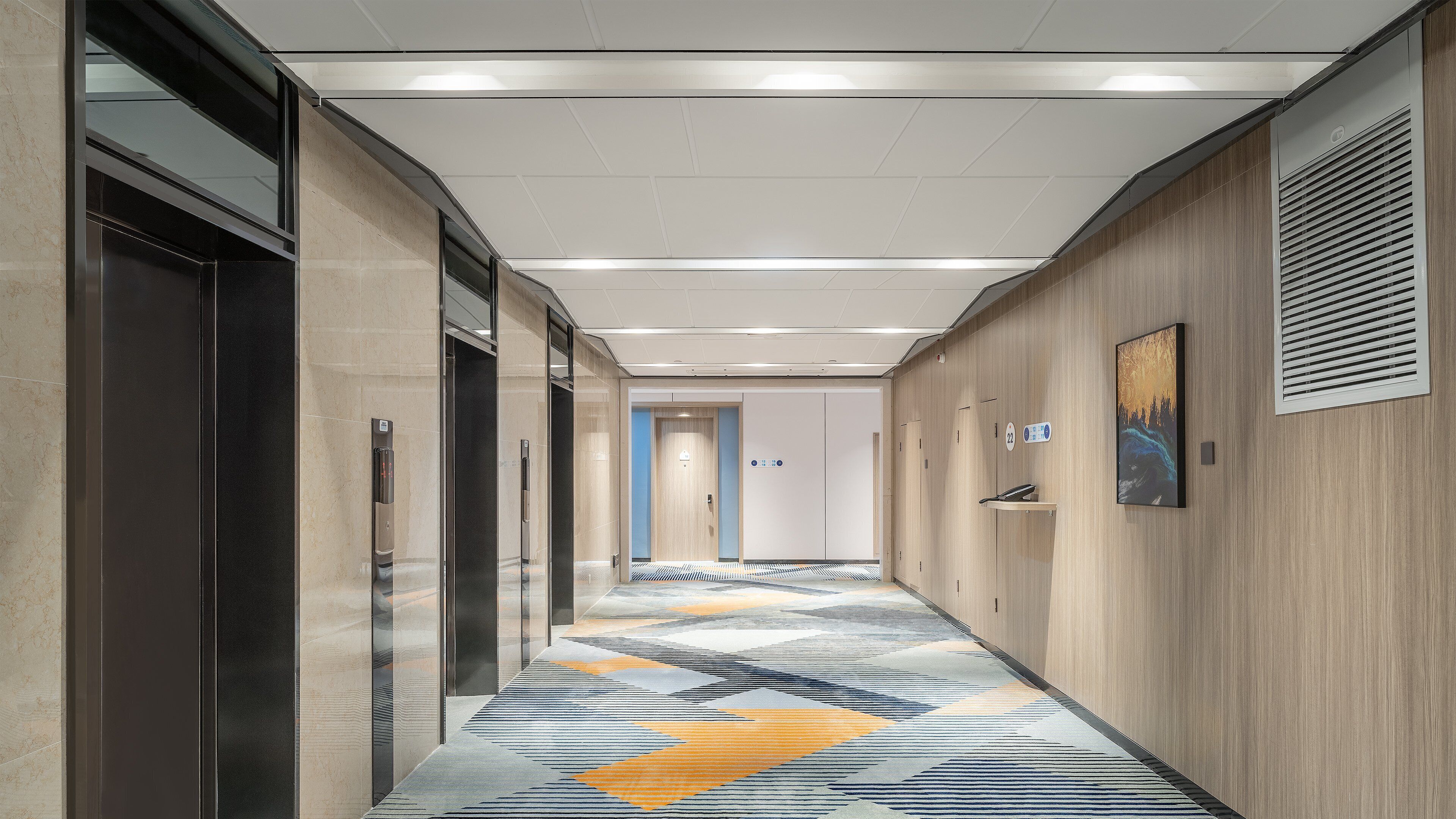 A modern hotel corridor with elevators, a patterned carpet, and a wooden wall with a painting.