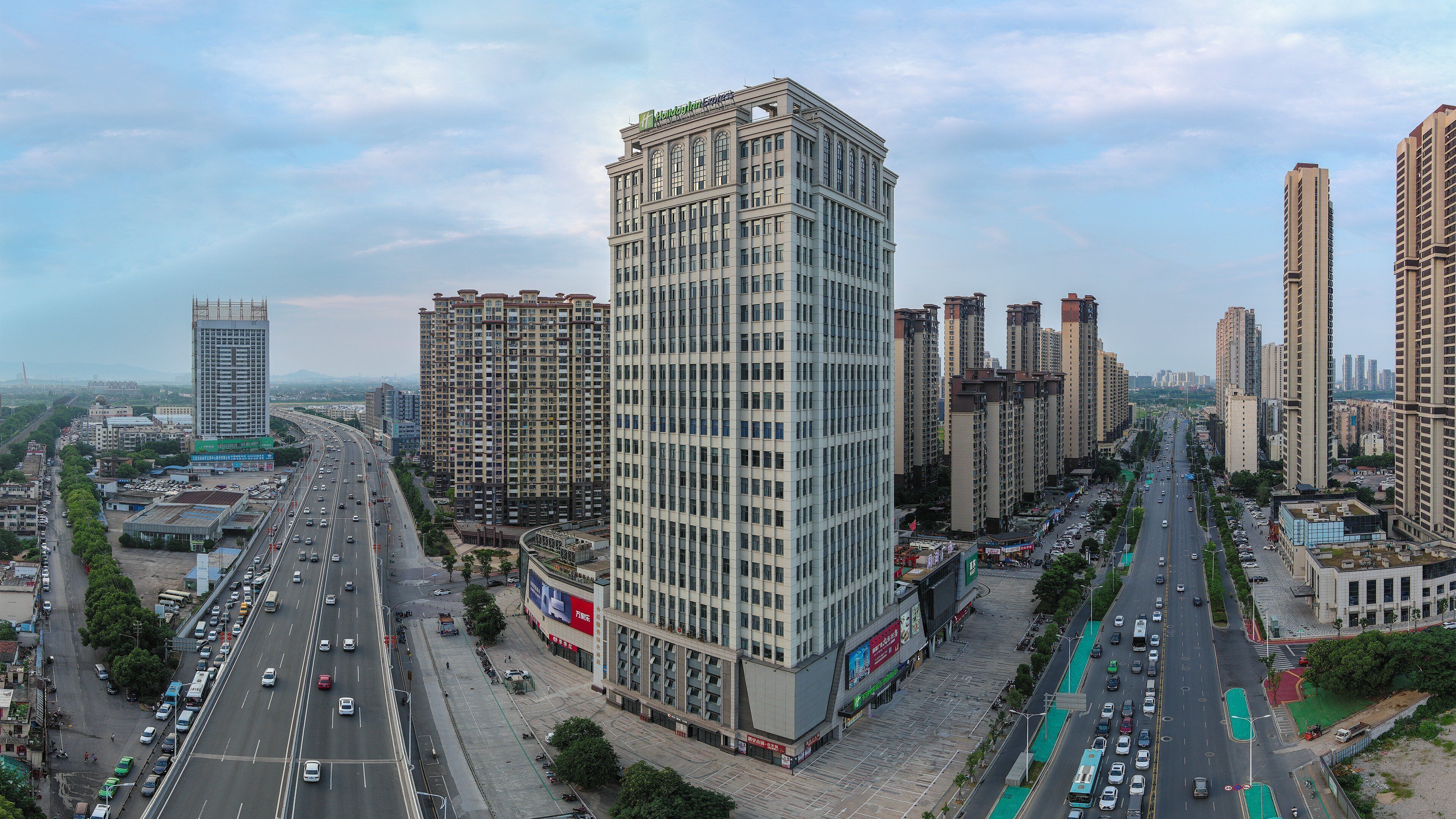 A high-rise building with multiple floors, surrounded by other tall buildings, a busy road with vehicles, and a clear sky.