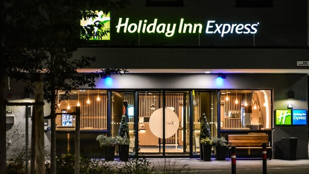 The exterior of a Holiday Inn Express hotel with a modern design, featuring a large glass entrance, outdoor seating, and a prominent logo.