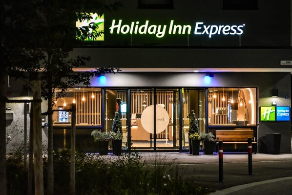 The exterior of a Holiday Inn Express hotel with a modern design, featuring a large glass entrance, outdoor seating, and a prominent logo. - Holiday Inn Express Munich Olching by IHG (Olching)