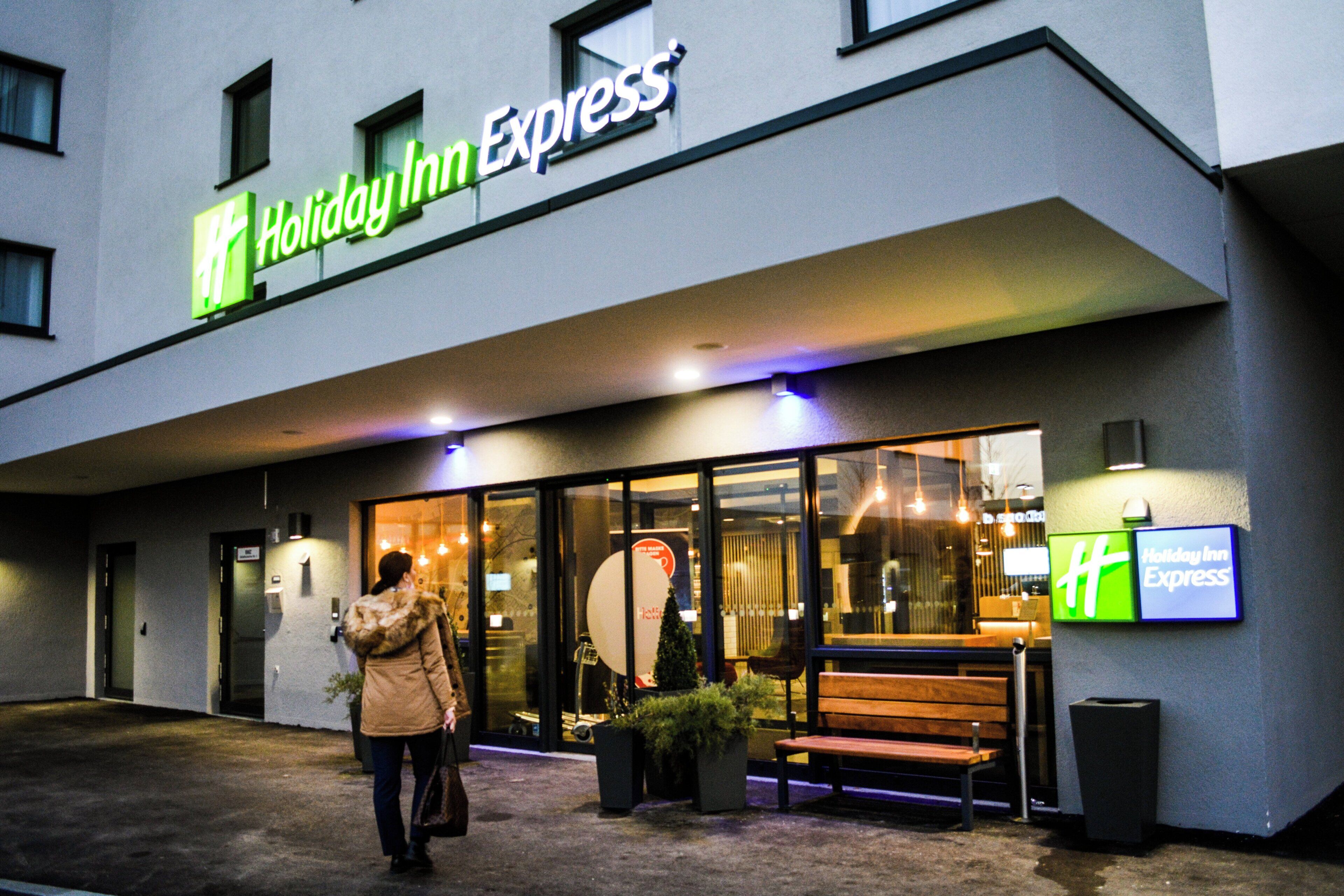 A Holiday Inn Express building with a glass entrance, a bench outside, and a person walking towards the entrance.