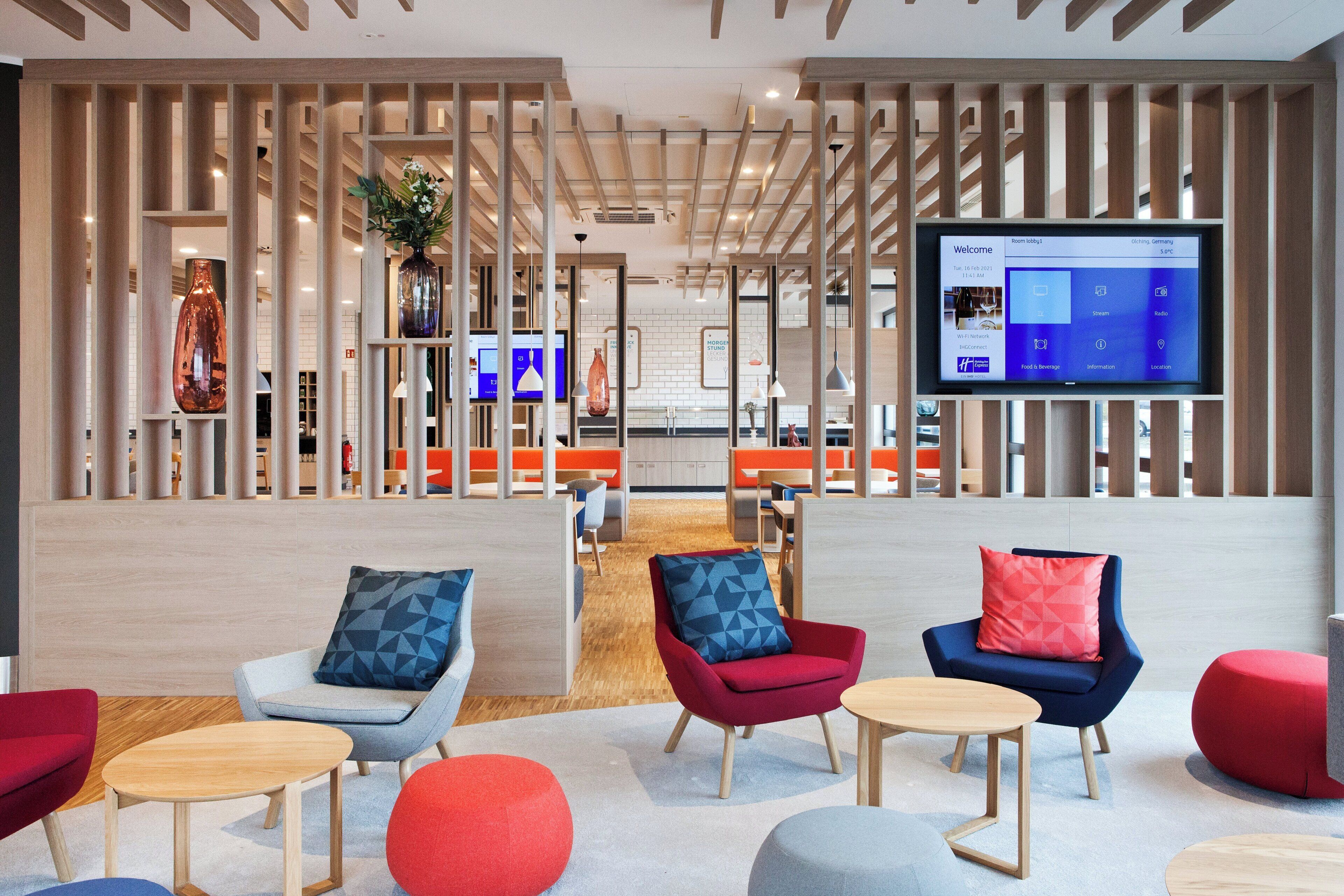 A modern hotel lobby with a reception desk, seating area with armchairs and ottomans, a large flat-screen TV displaying a welcome screen, and a wooden partition with a plant.