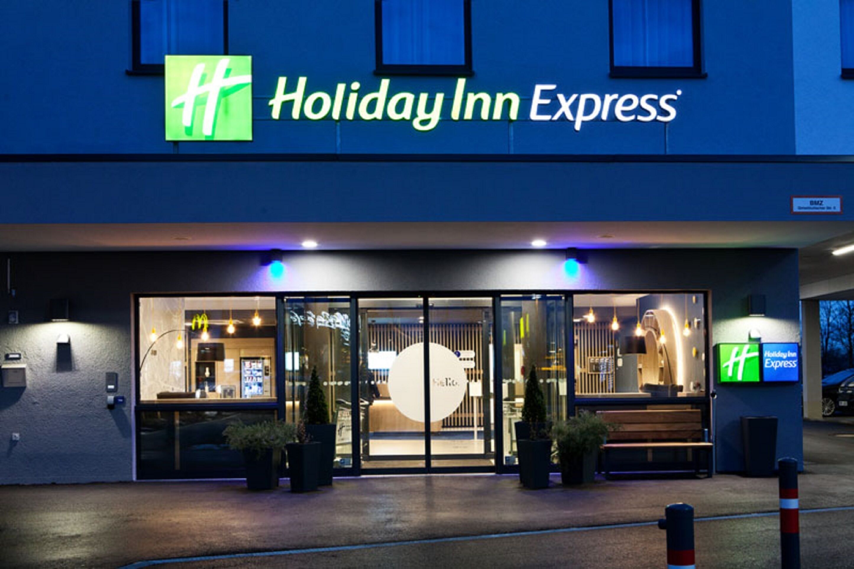 The exterior of a Holiday Inn Express hotel with a glass entrance, outdoor seating, and a visible sign.