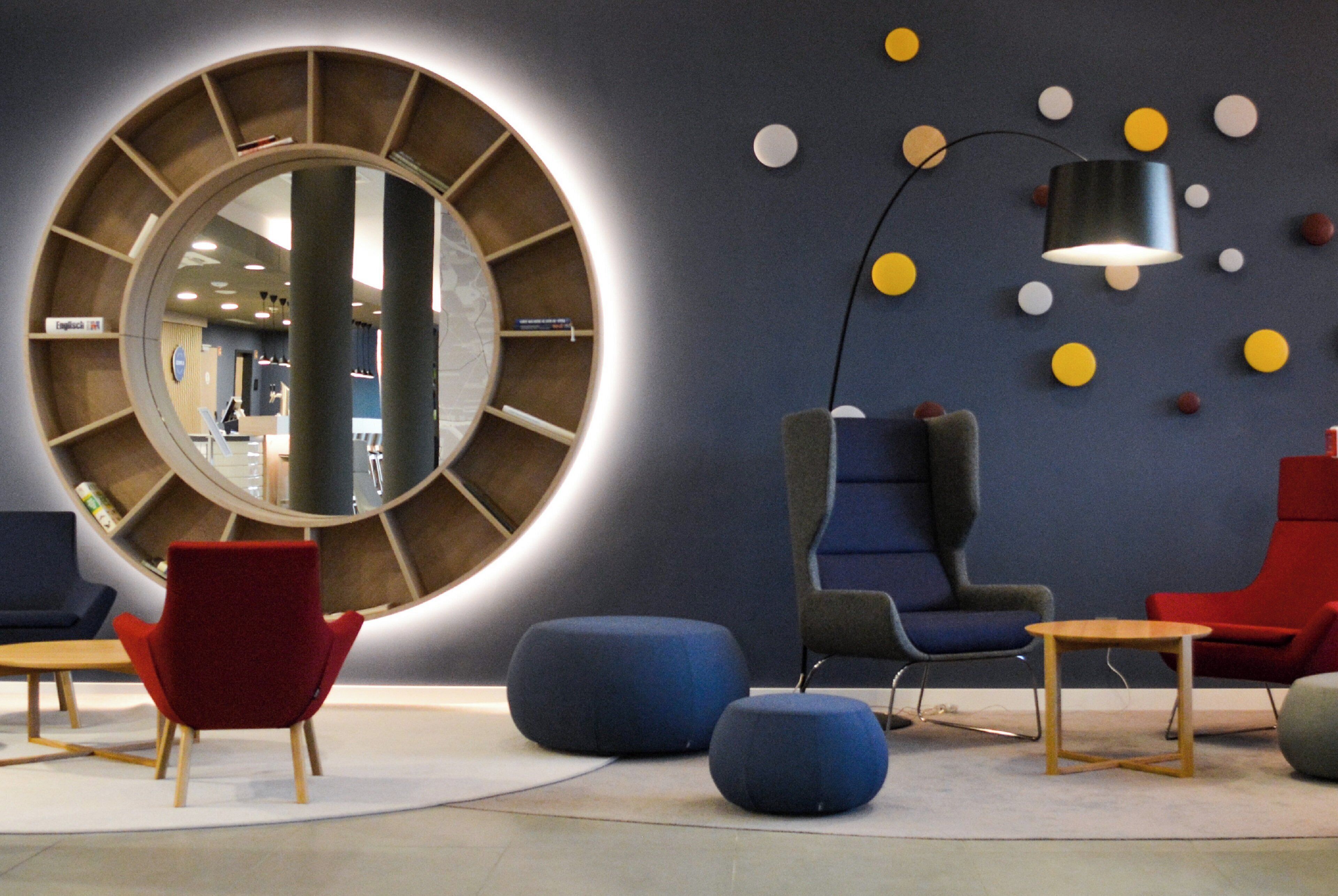 Ein moderner Loungebereich mit einem großen runden Spiegel, blauen und roten Sitzmöbeln sowie einer Stehlampe.