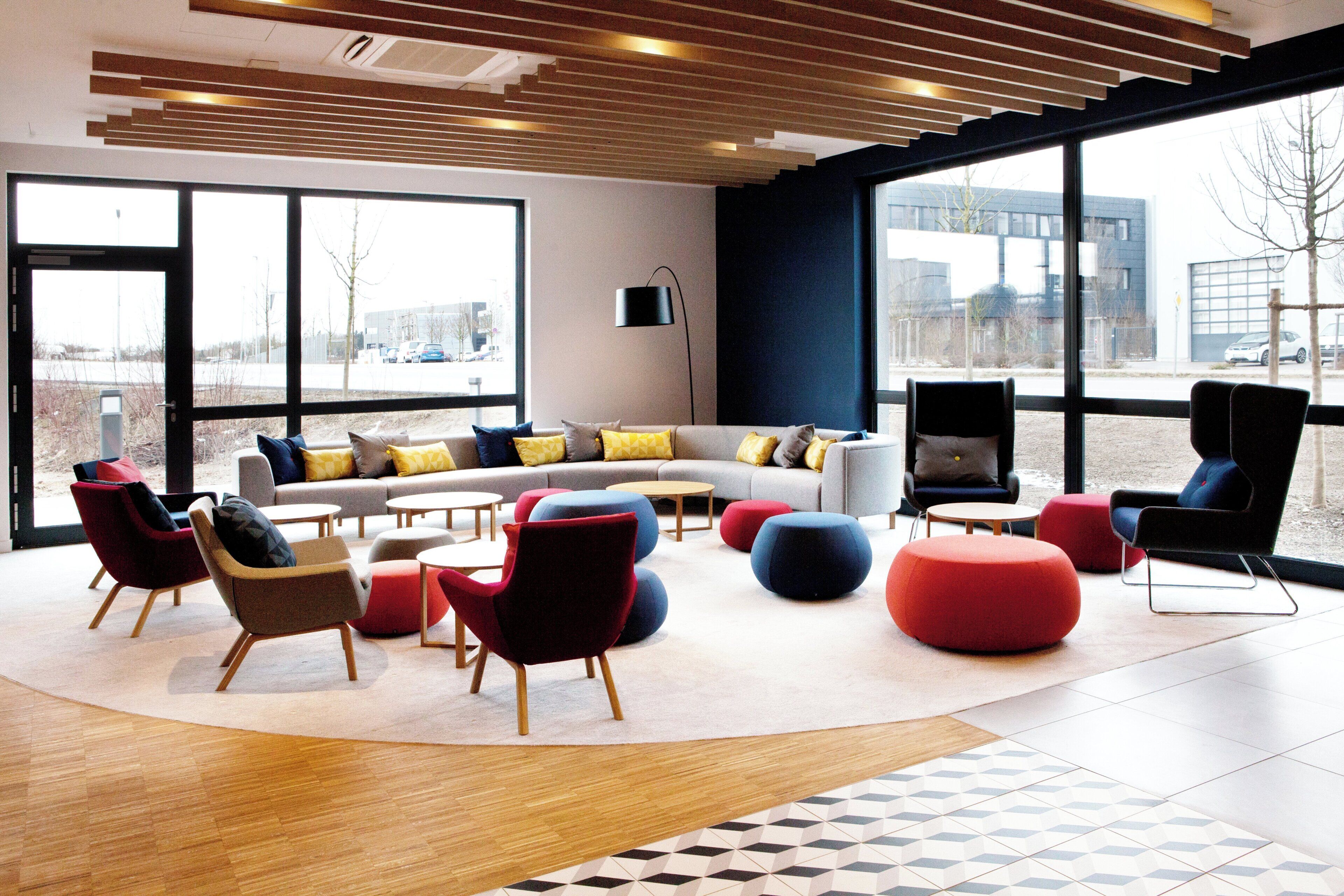 Een moderne loungehoek met een bank, stoelen, poefs en een groot raam met uitzicht op buiten.