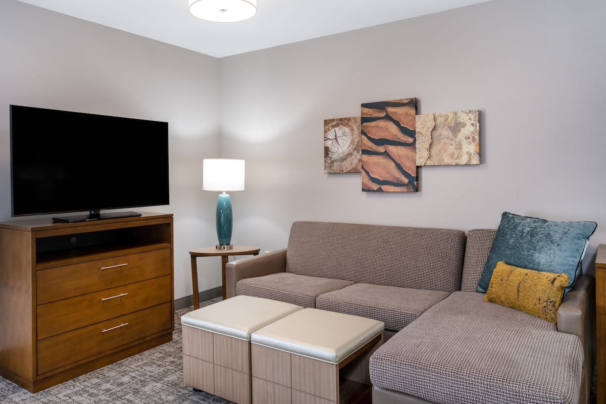A modern living room with a flat-screen TV, a wooden cabinet, a blue lamp, a sectional sofa with patterned cushions, and abstract wall art.