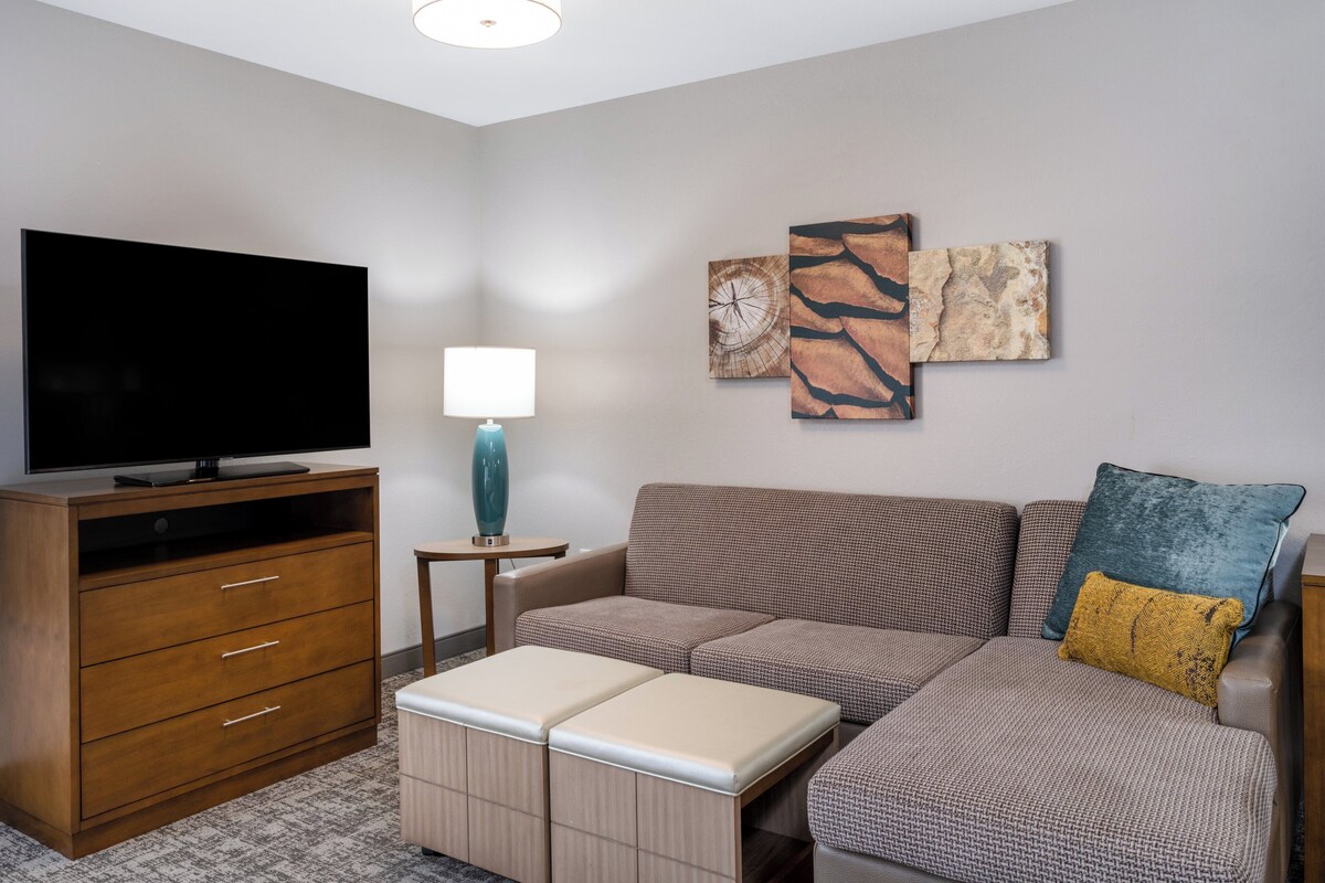 Una sala de estar moderna con un televisor de pantalla plana, un mueble de madera, una lámpara azul, un sofá modular con cojines estampados y una obra de arte abstracta en la pared.