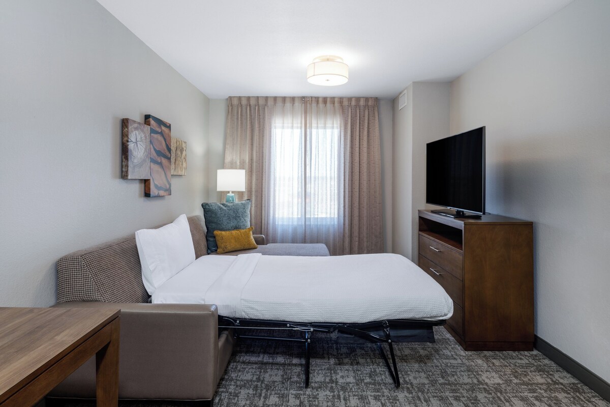 Una habitación de hotel con cama, escritorio, televisor y ventana con cortinas.