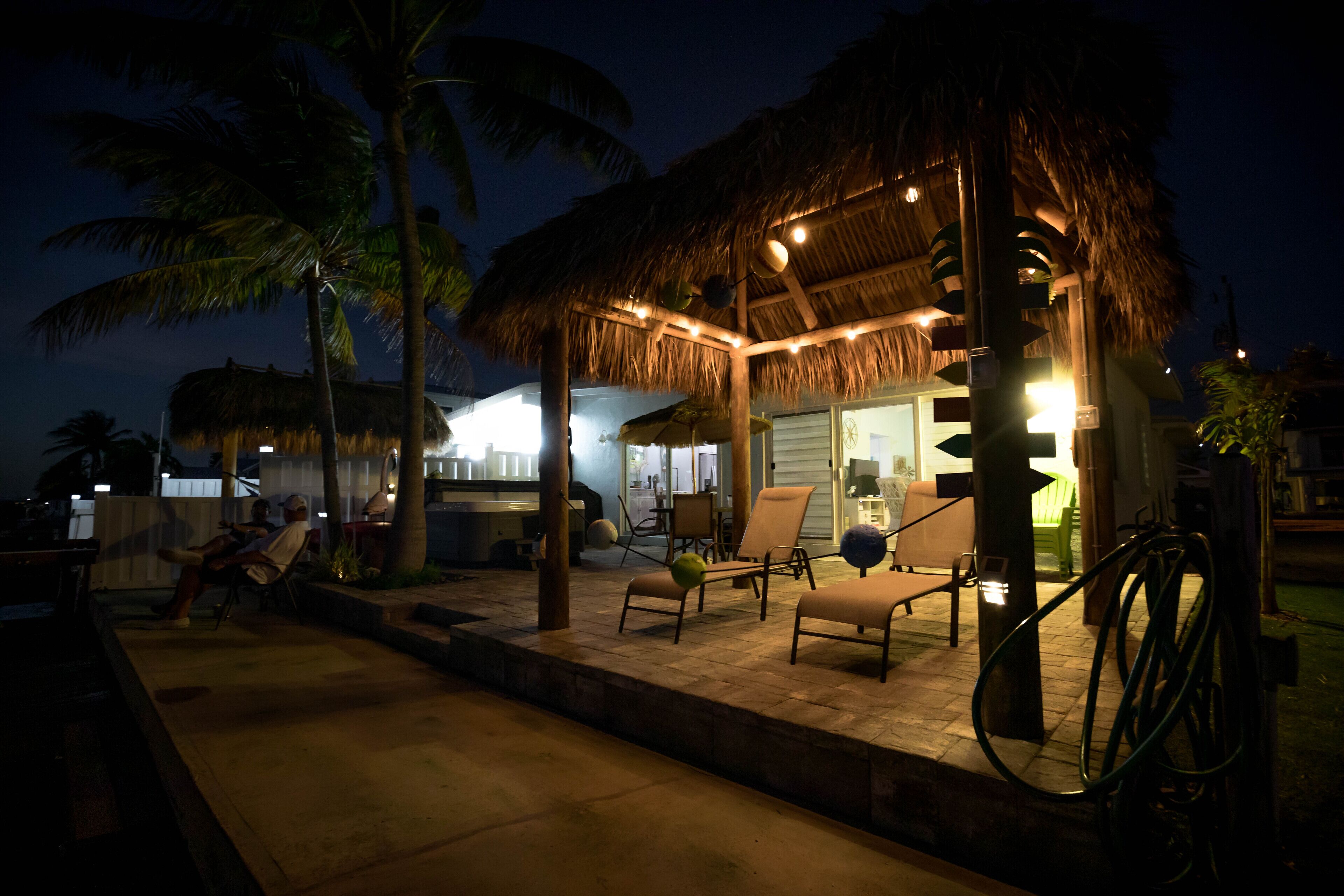 A thatched-roof outdoor area with lounge chairs and a table, surrounded by palm trees.