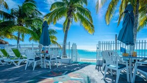 A beachside patio with white lounge chairs and tables, palm trees, and a view of the ocean. - ALL DOCKED OUT~ Key Colony Retreat Sandy beach's and sunsets (Key Colony Beach)