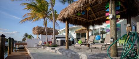 A wooden deck leading to a covered seating area with chairs and a table, surrounded by palm trees and a building with a thatched roof.
