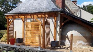 Una estructura de pérgola de madera con techo de listones, un muro de piedra y un sendero de piedra que la conduce.