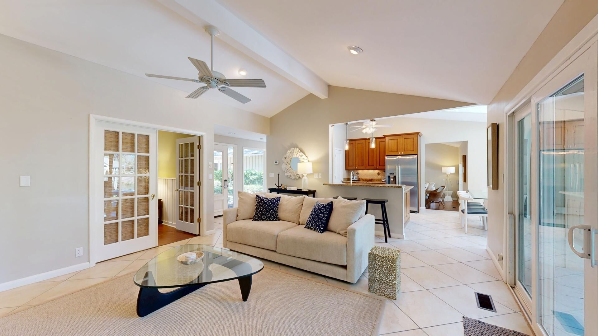A spacious living area with a glass coffee table, a beige sofa with patterned cushions, and a modern kitchen in the background.