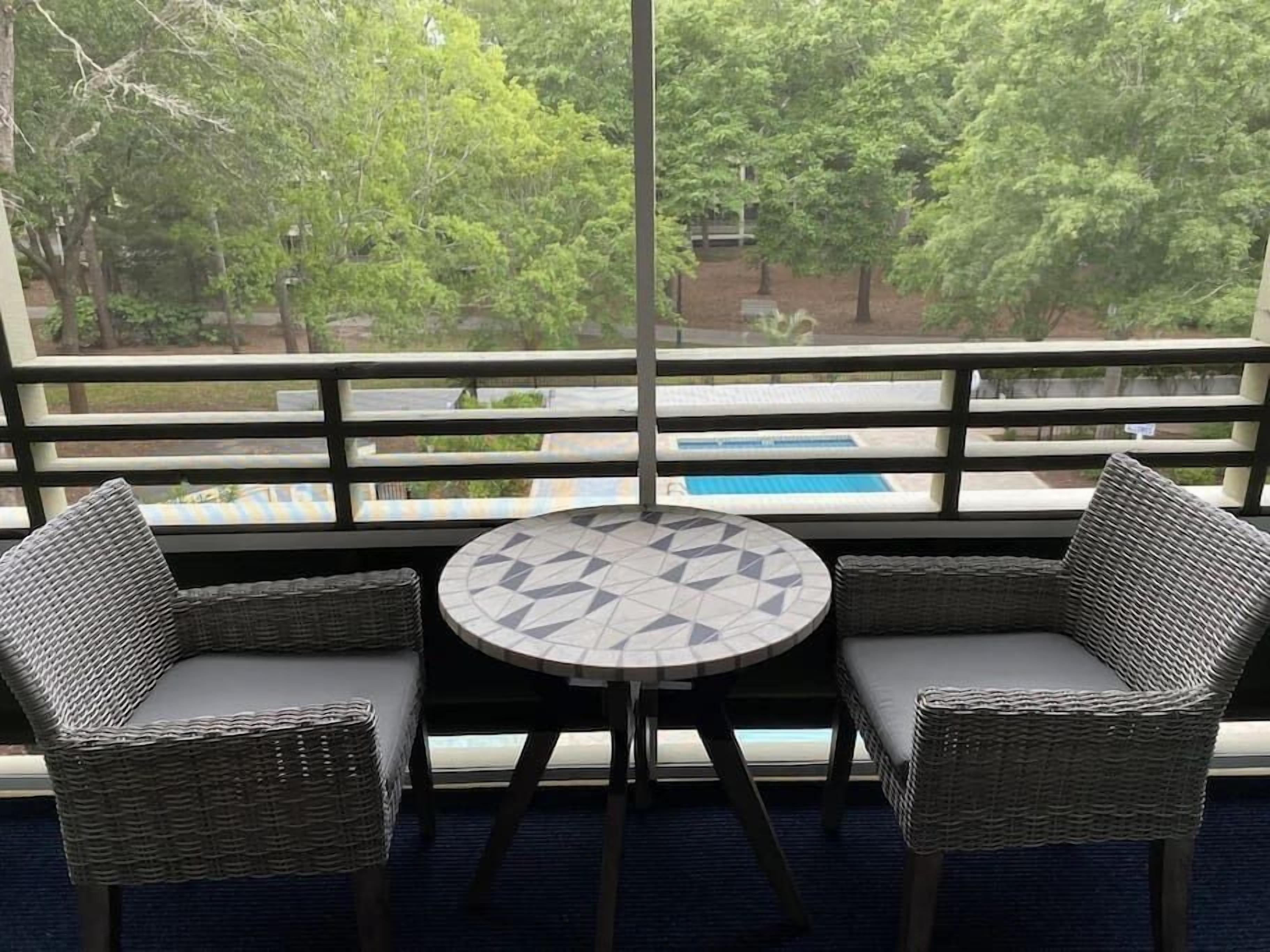 A balcony with wicker chairs and a table, overlooking a pool and greenery.
