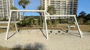 Un parque infantil con columpios y un arenero en una zona residencial con edificios altos al fondo.