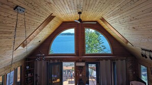 Eine Hütte mit schrägverlegtem Holzboden, einem großen Bogenfenster und Blick auf ein Gewässer.