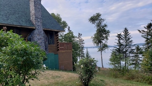 Eine Holzhütte mit einem Steinkamin, einer Holzterrasse und Blick auf einen von Bäumen umgebenen See.