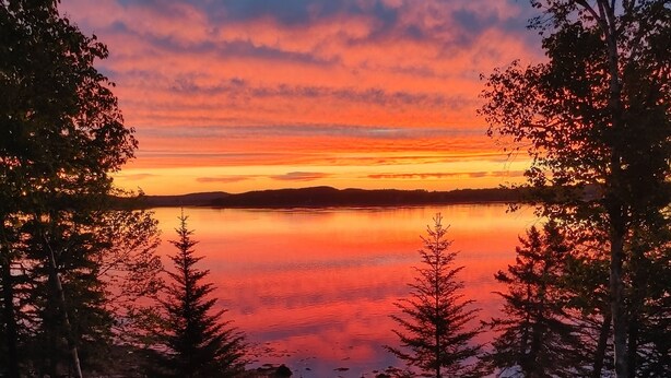 Blick auf einen See bei Sonnenuntergang, Bäume und eine Picknickplatz mit Stühlen und Feuerstelle.