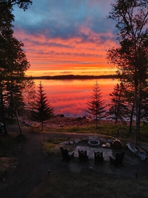 A lakeside view with a sunset, trees, and a picnic area with chairs and a fire pit. -   Waterfront log cabin/ Full refund up to 1 day out/Kayaks/ Fire pit/ beach (Lubec)