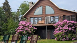 Eine zweistöckige Holzhütte mit Balkon, umgeben von üppigem Grün und blühenden Sträuchern.