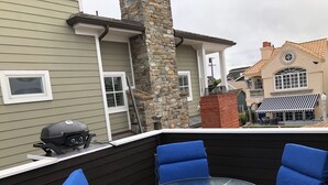 Un espacio de estar al aire libre techado con una mesa de vidrio y sillas azules acolchadas, una parrilla y un pilar de piedra.