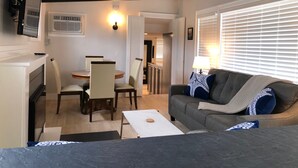 Una sala de estar acogedora con un sofá, una mesa de comedor con sillas, un televisor montado en la pared y una ventana con persianas.